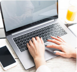 pessoa sentada digitando em um notebook com o celular ao lado que estão em cima de uma mesa.