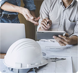 duas pessoas conversando próximas a uma mesa, e com objetvos espalhados em cima desta mesa: capacete de proteção para construçao civil, notebook e papéis.