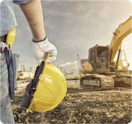 pessoa segurando com uma mão um capacete de proteção para construçao civil em um terreno com uma escavadeira em sua frente.